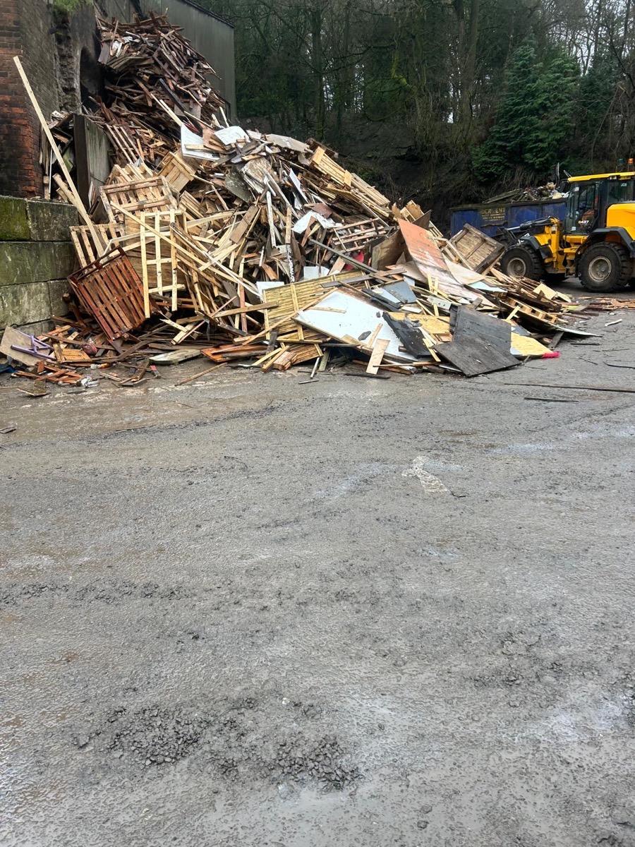 A JCB loader beside a large sorted pile of timber and pallets at the disposal site